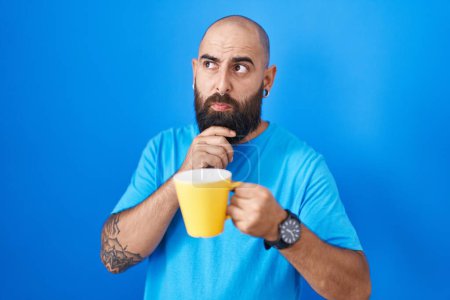 Téléchargez les photos : Jeune homme hispanique avec barbe et tatouages boire une tasse de café penser concentré sur le doute avec le doigt sur le menton et la recherche jusqu'à se demander - en image libre de droit