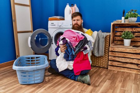 Redhead man with long beard putting dirty laundry into washing machine skeptic and nervous, frowning upset because of problem. negative person. 