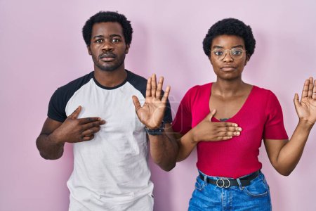 Foto de Joven pareja afroamericana de pie sobre fondo rosa jurando con la mano en el pecho y la palma abierta, haciendo un juramento de lealtad promesa - Imagen libre de derechos