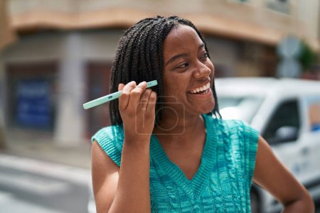Téléchargez les photos : Femme afro-américaine souriant confiant écoute message audio par le smartphone à la rue - en image libre de droit