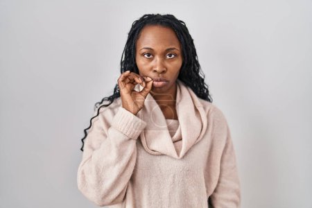 Téléchargez les photos : Femme africaine debout sur fond blanc bouche et lèvres fermées comme zip avec les doigts. secret et silencieux, tabou parlant - en image libre de droit