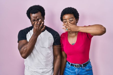 Foto de Joven pareja afroamericana de pie sobre fondo rosa oliendo algo apestoso y asqueroso, olor intolerable, conteniendo la respiración con los dedos en la nariz. mal olor - Imagen libre de derechos