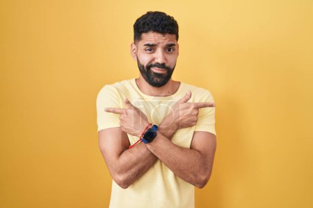 Foto de Hombre hispano con barba de pie sobre fondo amarillo apuntando a ambos lados con los dedos, en diferente dirección en desacuerdo - Imagen libre de derechos