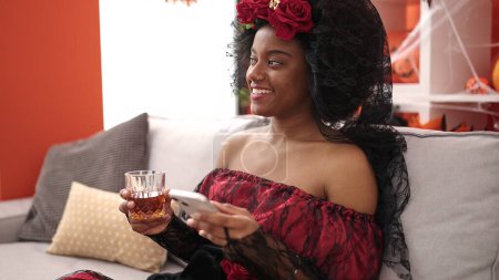 Téléchargez les photos : African american woman wearing katrina costume using smartphone drinking whisky at home - en image libre de droit