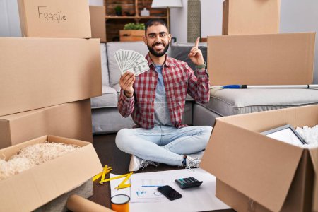 Photo for Middle east man with beard sitting on the floor at new home holding money surprised with an idea or question pointing finger with happy face, number one - Royalty Free Image