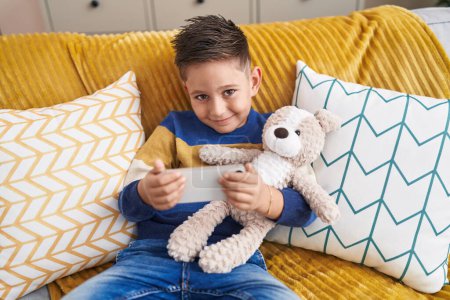 Photo for Adorable hispanic boy hugging teddy bear watching video on smartphone at home - Royalty Free Image
