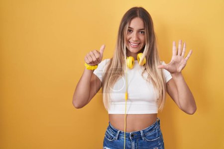 Foto de Mujer rubia joven de pie sobre fondo amarillo con auriculares que muestran y señalan hacia arriba con los dedos número seis mientras sonríe confiado y feliz. - Imagen libre de derechos