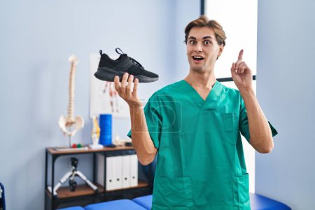 Photo for Young man working at physiotherapy clinic holding shoe surprised with an idea or question pointing finger with happy face, number one - Royalty Free Image