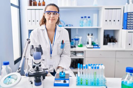 Téléchargez les photos : Jeune femme hispanique travaillant au laboratoire scientifique souriant regardant vers le côté et regardant loin en pensant. - en image libre de droit