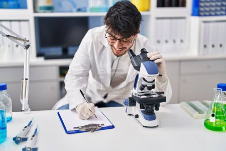Photo for Young hispanic man scientist writing on document using microscope at laboratory - Royalty Free Image