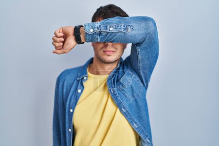 Photo for Young hispanic man standing over blue background covering eyes with arm, looking serious and sad. sightless, hiding and rejection concept - Royalty Free Image