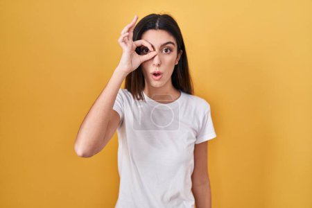 Foto de Young beautiful woman standing over yellow background doing ok gesture shocked with surprised face, eye looking through fingers. unbelieving expression. - Imagen libre de derechos