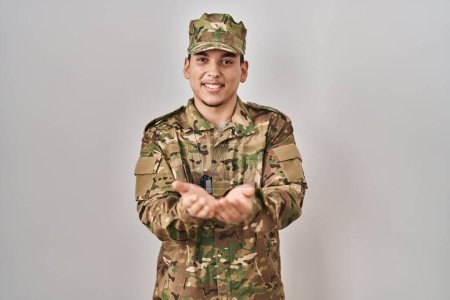 Foto de Joven árabe vestido con camuflaje uniforme del ejército sonriendo con las palmas de las manos juntas recibiendo o dando gesto. retención y protección - Imagen libre de derechos