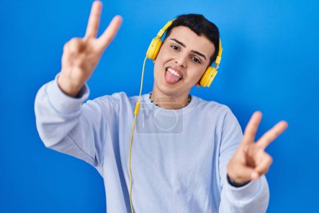 Photo for Non binary person listening to music using headphones smiling with tongue out showing fingers of both hands doing victory sign. number two. - Royalty Free Image