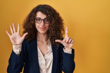 Photo for Hispanic woman with curly hair standing over yellow background showing and pointing up with fingers number seven while smiling confident and happy. - Royalty Free Image