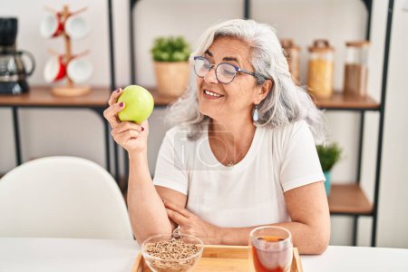 Téléchargez les photos : Femme d'âge moyen souriant confiant tenant pomme à la maison - en image libre de droit