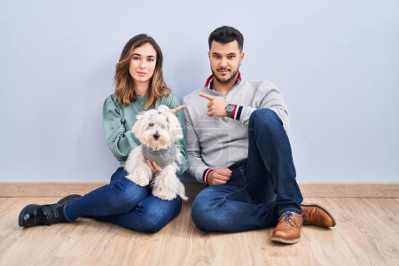 Foto de Young hispanic couple sitting on the floor with dog pointing with hand finger to the side showing advertisement, serious and calm face - Imagen libre de derechos