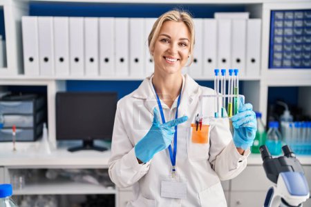 Téléchargez les photos : Jeune femme caucasienne travaillant au laboratoire scientifique tenant des éprouvettes souriant heureux pointant avec la main et le doigt - en image libre de droit
