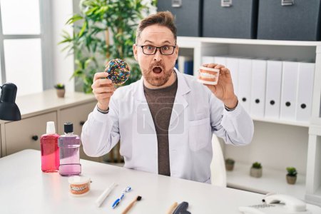 Photo for Middle age caucasian dentist man holding denture and doughnuts in shock face, looking skeptical and sarcastic, surprised with open mouth - Royalty Free Image