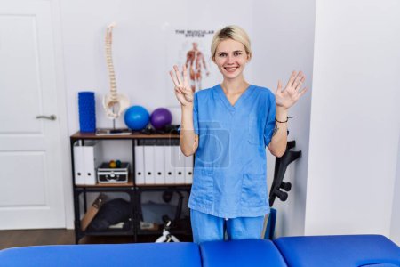 Foto de Young physiotherapist woman working at pain recovery clinic showing and pointing up with fingers number eight while smiling confident and happy. - Imagen libre de derechos