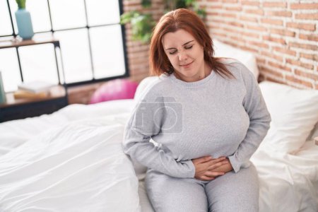 Téléchargez les photos : Jeune belle femme plus la taille souffrant de douleur menstruelle assis sur le lit dans la chambre - en image libre de droit