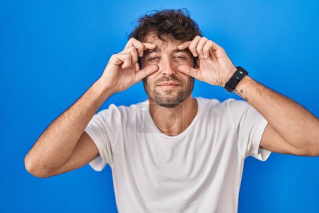 Téléchargez les photos : Hispanic young man standing over blue background trying to open eyes with fingers, sleepy and tired for morning fatigue - en image libre de droit
