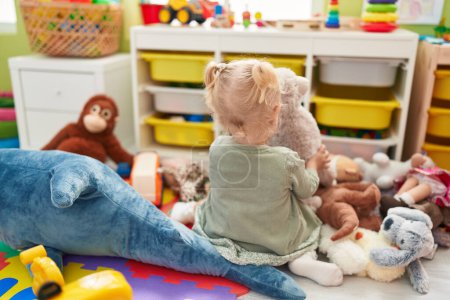 Foto de Adorable chica rubia jugando con oso de peluche sentado en el suelo en el jardín de infantes - Imagen libre de derechos