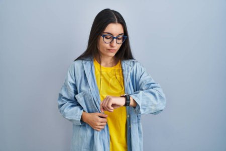 Foto de Mujer hispana joven de pie sobre fondo azul comprobando la hora en el reloj de pulsera, relajada y confiada - Imagen libre de derechos