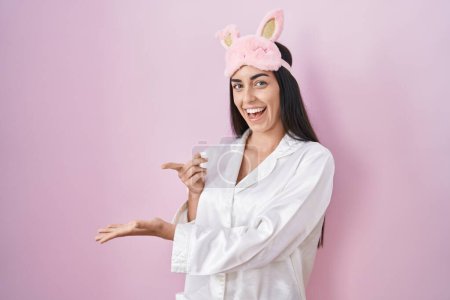 Téléchargez les photos : Young brunette woman wearing sleep mask and pajama amazed and smiling to the camera while presenting with hand and pointing with finger. - en image libre de droit