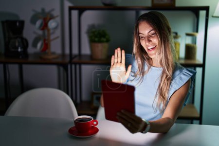Téléchargez les photos : Jeune femme hispanique utilisant touchpad assis sur la table la nuit renonçant à dire bonjour heureux et souriant, geste de bienvenue convivial - en image libre de droit