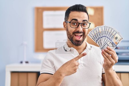 Téléchargez les photos : Jeune homme hispanique avec barbe et lunettes tenant des billets en dollars souriant heureux pointant avec la main et le doigt - en image libre de droit