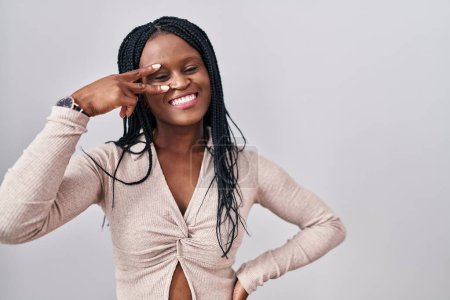 Téléchargez les photos : Femme africaine avec des tresses debout sur fond blanc faisant symbole de paix avec les doigts sur le visage, souriant joyeuse montrant la victoire - en image libre de droit