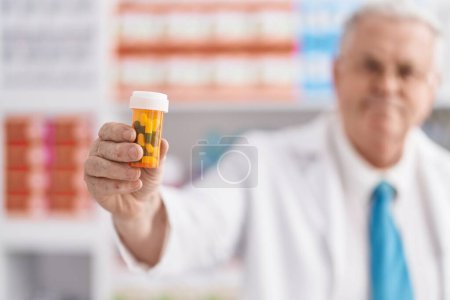 Foto de Middle age grey-haired man pharmacist smiling confident holding pills bottle at pharmacy - Imagen libre de derechos