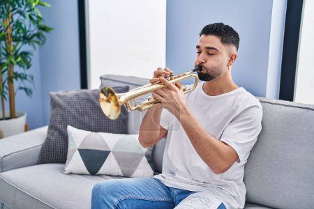 Téléchargez les photos : Jeune homme arabe musicien jouer de la trompette assis sur le canapé à la maison - en image libre de droit
