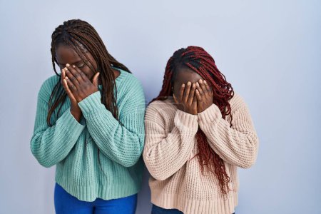 Foto de Dos mujeres africanas de pie sobre fondo azul con expresión triste cubriendo la cara con las manos mientras lloran. concepto de depresión. - Imagen libre de derechos