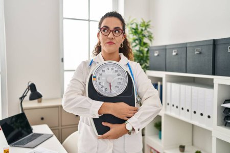 Téléchargez les photos : Jeune femme hispanique travaillant à la clinique diététiste souriant regardant vers le côté et regardant loin en pensant. - en image libre de droit
