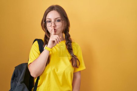 Téléchargez les photos : Young caucasian woman wearing student backpack over yellow background asking to be quiet with finger on lips. silence and secret concept. - en image libre de droit