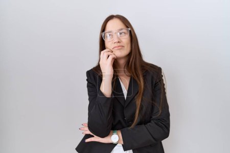 Foto de Beautiful brunette woman wearing business jacket and glasses with hand on chin thinking about question, pensive expression. smiling with thoughtful face. doubt concept. - Imagen libre de derechos