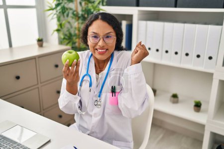 Photo for Young hispanic doctor woman holding green apple screaming proud, celebrating victory and success very excited with raised arm - Royalty Free Image