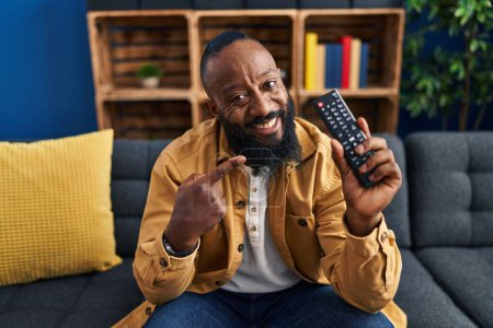 Foto de African american man holding television remote control smiling happy pointing with hand and finger - Imagen libre de derechos