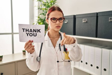 Téléchargez les photos : Young caucasian woman wearing doctor uniform holding thank you banner pointing with finger to the camera and to you, confident gesture looking serious - en image libre de droit