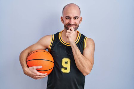 Téléchargez les photos : Young bald man with beard wearing basketball uniform holding ball looking confident at the camera smiling with crossed arms and hand raised on chin. thinking positive. - en image libre de droit