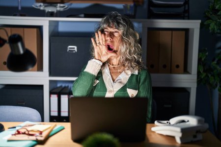 Foto de Middle age woman working at night using computer laptop shouting and screaming loud to side with hand on mouth. communication concept. - Imagen libre de derechos