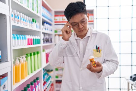 Téléchargez les photos : Young chinese man pharmacist looking pills bottle at pharmacy - en image libre de droit