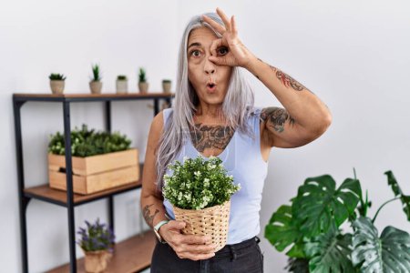 Téléchargez les photos : Femme aux cheveux gris du moyen âge tenant pot de plante verte à la maison faisant un geste correct choqué par le visage surpris, les yeux regardant à travers les doigts. expression incrédule. - en image libre de droit