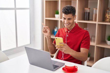 Photo for Young hispanic man using laptop and doing video call with smartphone smiling happy pointing with hand and finger to the side - Royalty Free Image