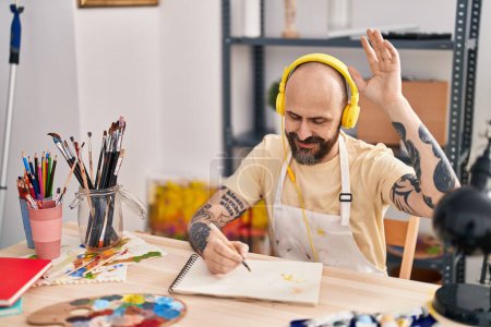 Téléchargez les photos : Jeune homme chauve artiste écoutant de la musique dessiner sur carnet au studio d'art - en image libre de droit