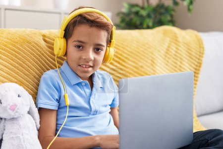 Photo for Adorable hispanic boy using laptop and headphones sitting on sofa at home - Royalty Free Image