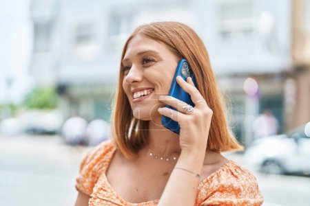 Téléchargez les photos : Jeune rousse femme souriant confiant parler sur le smartphone à la rue - en image libre de droit