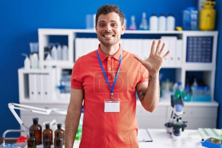 Photo for Young hispanic man working at scientist laboratory showing and pointing up with fingers number five while smiling confident and happy. - Royalty Free Image